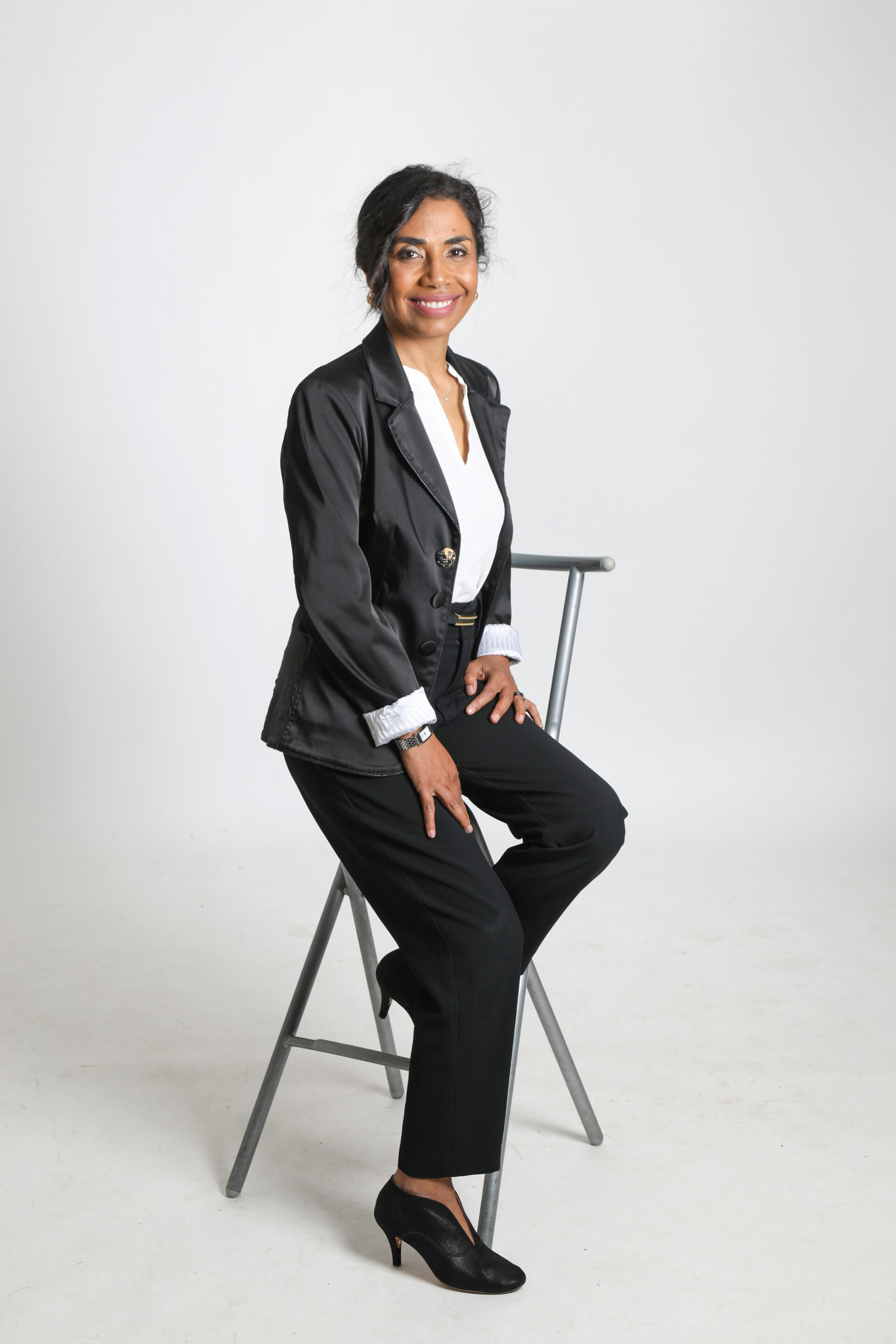 Portrait en pied du Dr Joéline Andriana, Docteur en Psychologie Sociale. Elle est assise sur un tabouret haut, vêtue d'un tailleur noir élégant, illustrant son professionnalisme et son expertise scientifique pour sa page dédiée à son parcours de psychologue et de chercheuse.